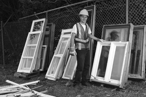 A male worker wearing a safety helmet and high-visibility vest stands outdoors next to a collection of empty white window frames with glass panes, arranged upright and leaning against a chain-link fence. The window frames, made of UPVC or timber, are of various sizes and are positioned in front of a natural background with trees and shrubbery. The ground is grassy with some loose parts of wood or frame components scattered nearby. The worker appears to be involved in the collection or disposal of old or surplus window units, relevant to commercial waste removal services in Covent Garden. The scene is captured in natural daylight, emphasizing the utilitarian environment suitable for rubbish removal tasks, with the focus on the frames and the worker preparing for collection or recycling.