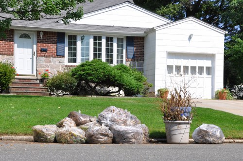A suburban front garden scene with a row of large, irregularly shaped rocks placed along the curb of a residential street. The rocks vary in size and color, with some displaying shades of gray, beige, and brown, and have rough, textured surfaces. Behind the rocks, a well-maintained green lawn extends towards a brick and white-panelled house with a prominent bay window and a small set of brick steps leading to a white front door. To the right of the rocks, a large white pot contains a dry, leafless shrub, and a smaller plant in a separate container is nearby. The house features a white garage door and is surrounded by trees and shrubs, with sunlight casting shadows indicating clear weather. The scene reflects routine disposal or removal of garden or household waste materials in a residential area, aligned with rubbish removal services in Covent Garden or similar districts.