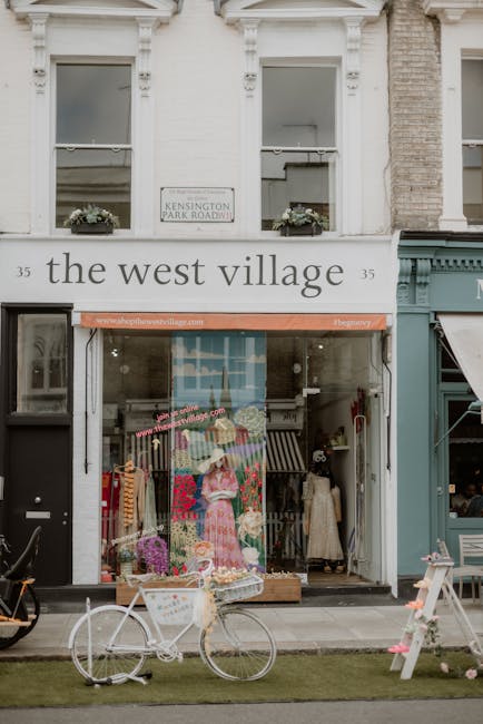 The storefront of a small retail shop named 'the west village' is visible, with a white facade and large glass windows displaying mannequins dressed in colorful clothing and floral arrangements. Above