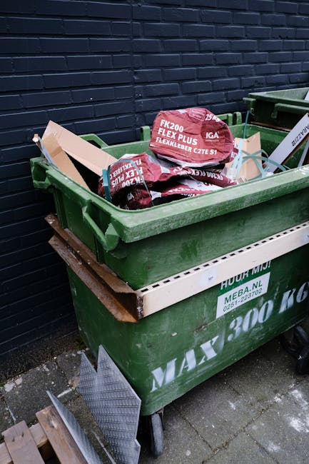 A narrow urban alleyway filled with a large collection of mixed waste, including black and grey recycling bags, cardboard boxes, and discarded packaging materials stacked behind a metal barricade. In 