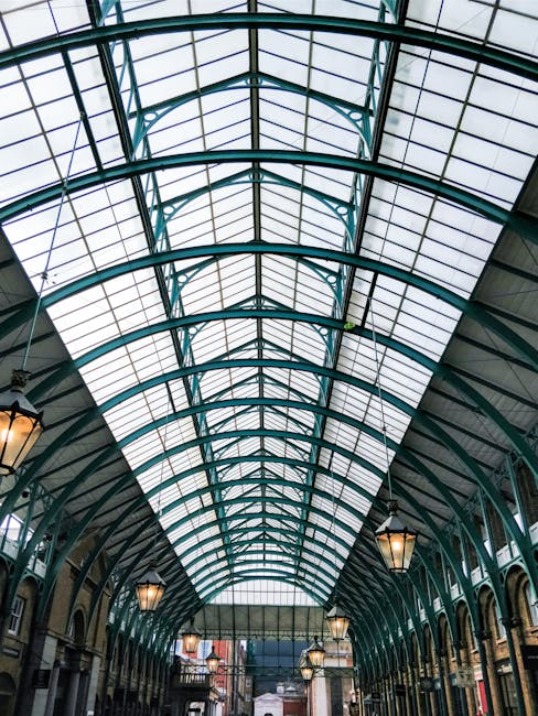 A large, curved glass and metal roof structure with a series of arched, teal-painted metal frames supporting rectangular glass panes, allowing natural light to filter through. In the background, there