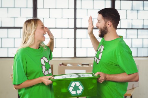 A photograph showing two ethnically diverse waste management professionals wearing bright green T-shirts with the recycling symbol, standing indoors in a well-lit industrial or warehouse setting. The woman on the left has long blonde hair and is smiling broadly, holding a small grey object in her right hand, while the man on the right has short dark hair and a beard, and is also smiling, giving a high-five with his right hand. Both are positioned towards each other, with the man holding a transparent green recycling bin in his left hand, featuring the recycling symbol on its front. The background includes large windows with grid-like frames, allowing natural light to illuminate the scene, and a stack of cardboard boxes can be seen behind them. The setting and their attire suggest they are engaged in waste disposal or recycling activities, consistent with professional rubbish removal services within the Covent Garden area.